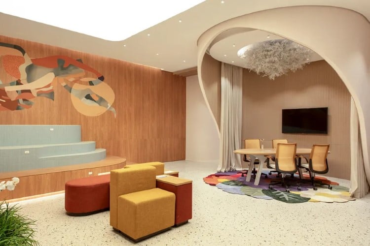 A imagem mostra um interior moderno e elegante com uma área de estar e jantar. Destacam-se sofás geométricos coloridos, mesa de jantar com cadeiras, e decoração artística nas paredes de madeira.