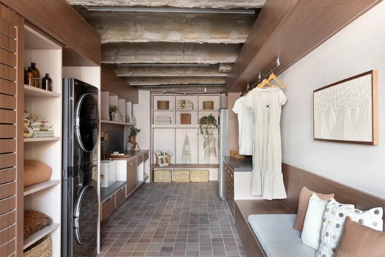 A imagem mostra um interior de casa compacta e bem organizada, com área de lavar roupa. Há prateleiras com utensílios, quadros decorativos na parede, e uma área de estar com sofá.