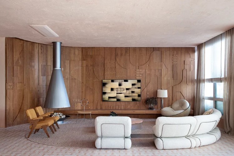 A sala de estar moderna tem paredes e piso em tons neutros e madeira, sofá branco curvo, poltronas de couro, lareira cônica suspensa, mesas de centro baixas e obra de arte geométrica. Janelas trazem luz natural.
