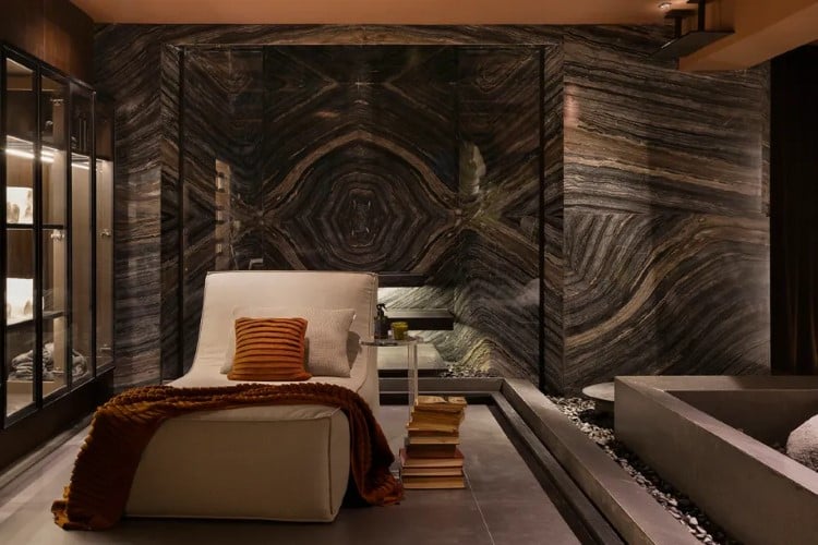 A imagem exibe um quarto elegante com paredes e piso de mármore escuro e padrões únicos. Há uma cama com cabeceira branca, cobertores marrons e almofadas laranjas, ao lado de uma mesa de vidro.