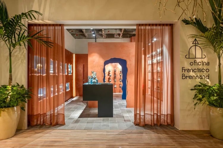 A entrada da galeria “Oficina Francisco Brennand” é emoldurada por cortinas laranjas, com plantas verdes ao lado. Dentro, há esculturas e uma parede texturizada ao fundo.