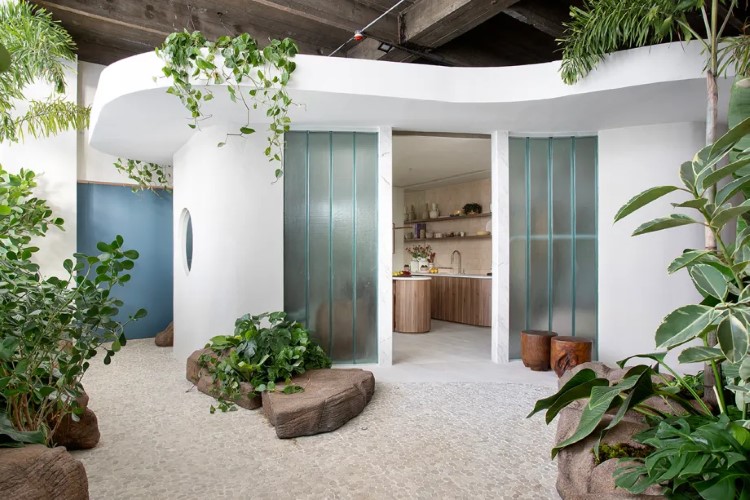A imagem mostra um interior moderno com paredes curvas brancas, piso de pedra, plantas verdes, móveis de madeira e portas de vidro.