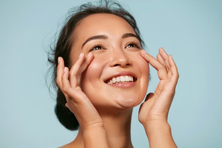 Mulher asiática com pele sem acnes e poros, sorrindo e passando mão no rosto