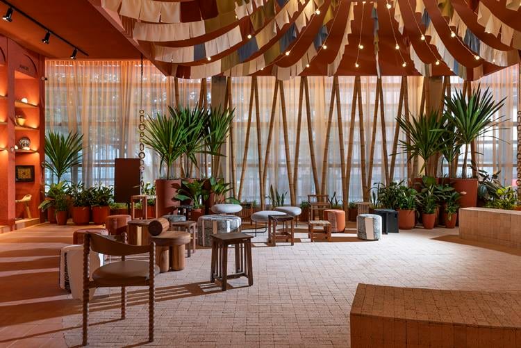 A imagem mostra um interior moderno e estiloso, com decoração tropical. Inclui cadeiras e mesas contemporâneas, plantas verdes, iluminação suave e teto com fitas de madeira curvas.