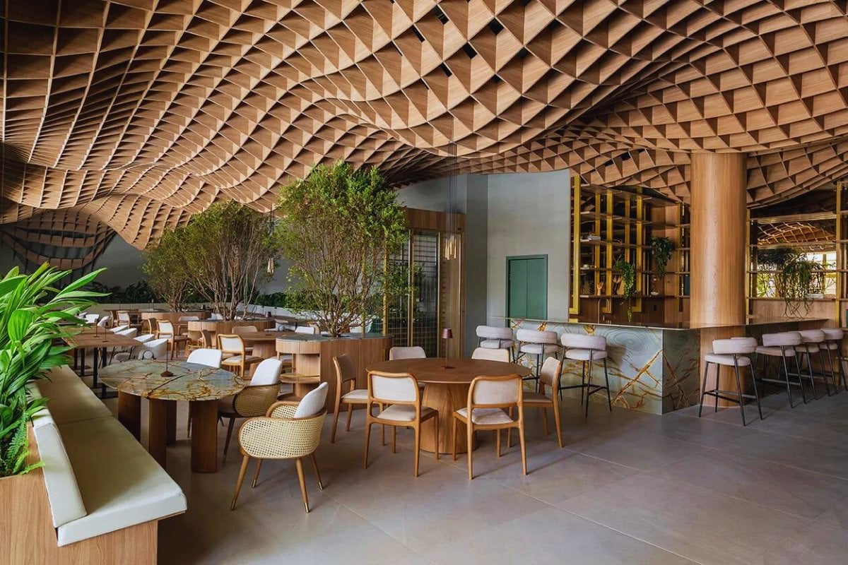 A imagem mostra um espaço interior moderno e elegante, com um teto ondulado de madeira que cria um padrão geométrico interessante. Mobiliário de madeira clara e plantas verdes complementam o ambiente.