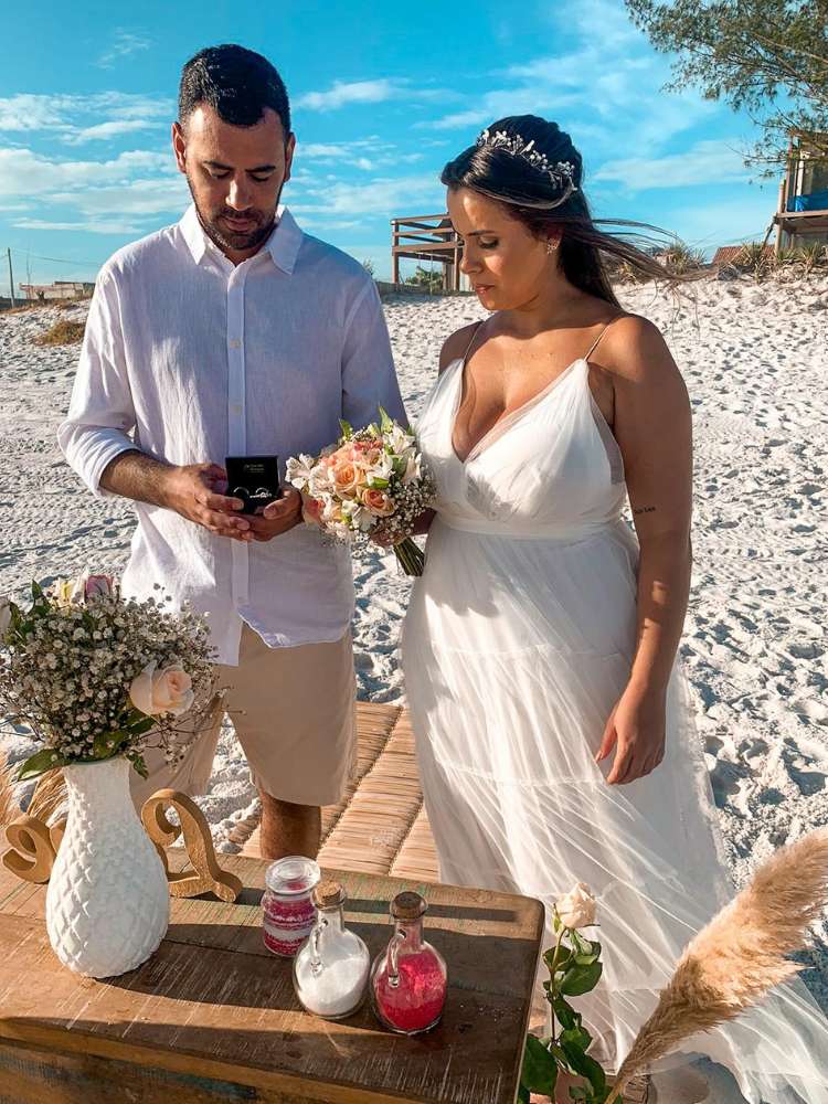 Noivo e noiva em casamento na praia com altar decorado com vaso branco e garrafas de vidro com areia colorida em vermelho e branco