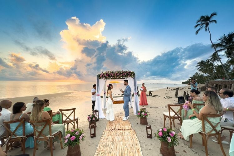 Casal celebrando casamento na praia, com mar ao fundo, convidados sentados e tapete de palha até o "altar"