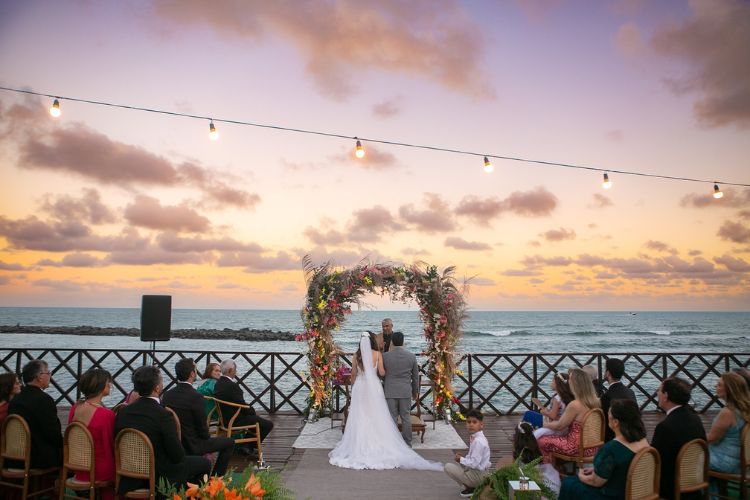 Pôr do sol com casamento na praia, noivos, arco floral e convidados