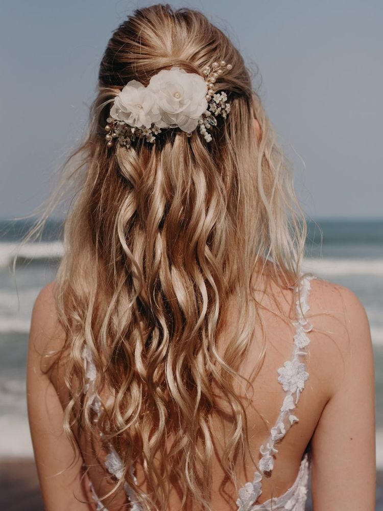 Mulher loira de pele clara de costas para a foto e de frente para o mar usando cabelo solto com presilhas de flor