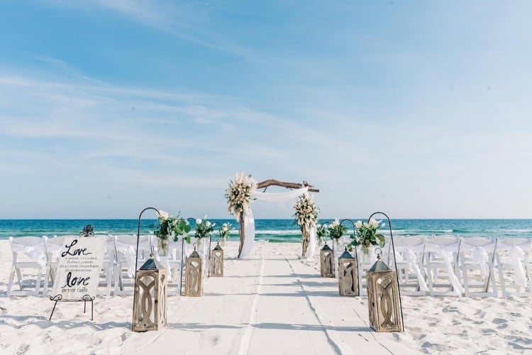 Casamento na praia com céu azul, cadeiras brancas, altar com flores e decoração clean