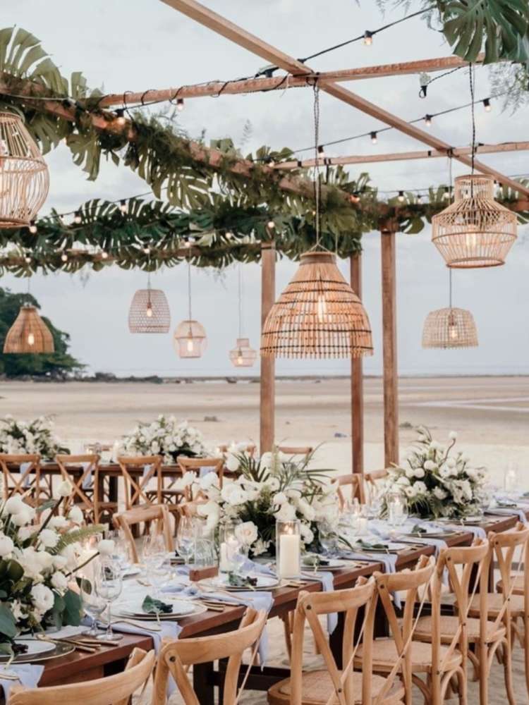 Mesas decoradas na praia com flores claras, velas na mesa, plantas na estrutura e lustres