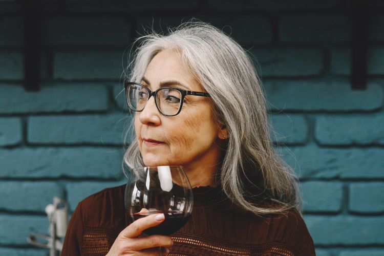 Mulher com cabelo grisalho de roupa marrom com taça de vinho na mão usando óculos de grau