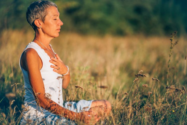 Mulher de cabelo curto com roupa branca meditando sentada em mato