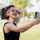 Mulher de pele clara usando regata preta, tirando selfie segurando cabelo curto cacheado com mechas rosas