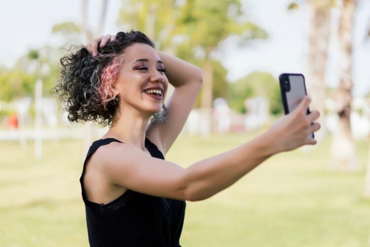 Mulher de pele clara usando regata preta, tirando selfie segurando cabelo curto cacheado com mechas rosas