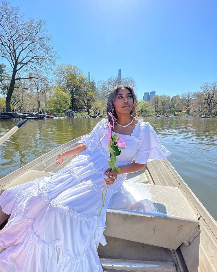 Mulher sentada em barco usando vestido branco com babaos e segurando rosa