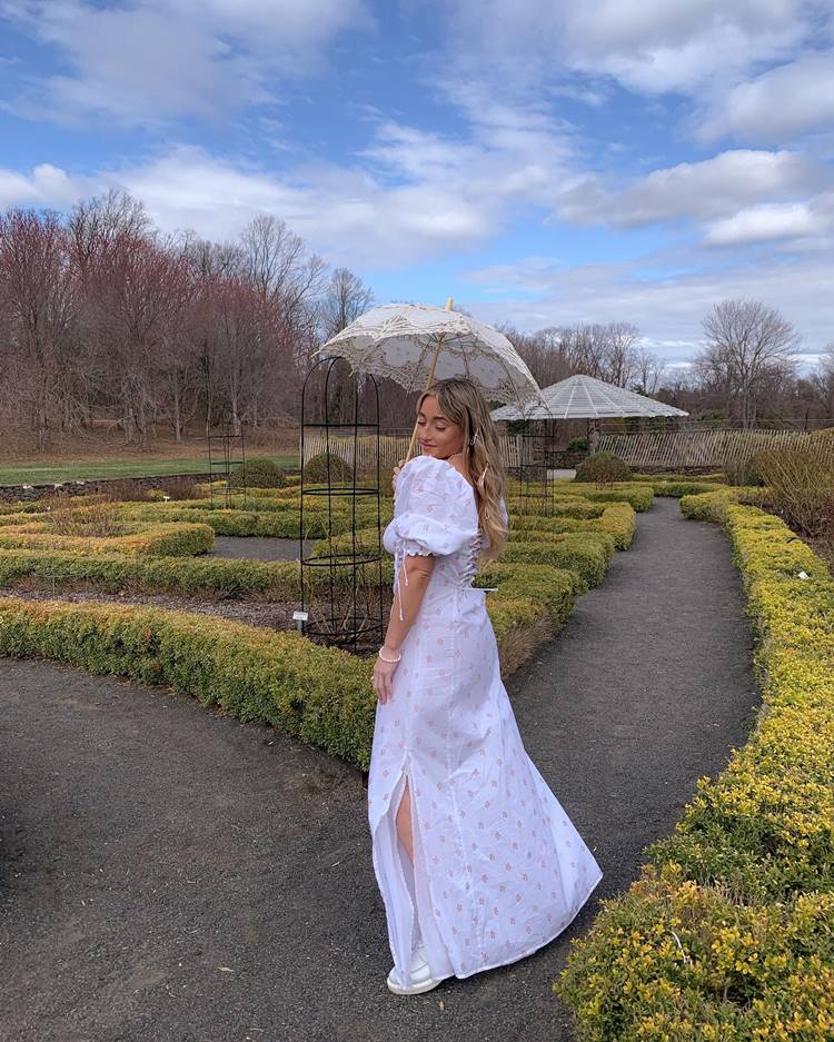 Mulher usando vestido com amarração nas costas, tênis e sombrinha de renda em um parque