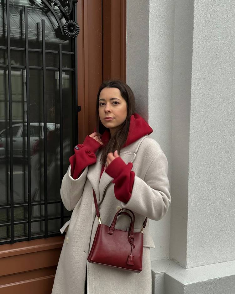 Mulher com casaco cinza-claro com moletom vermelho-cereja por baixo + bolsa da mesma cor, encostada em porta