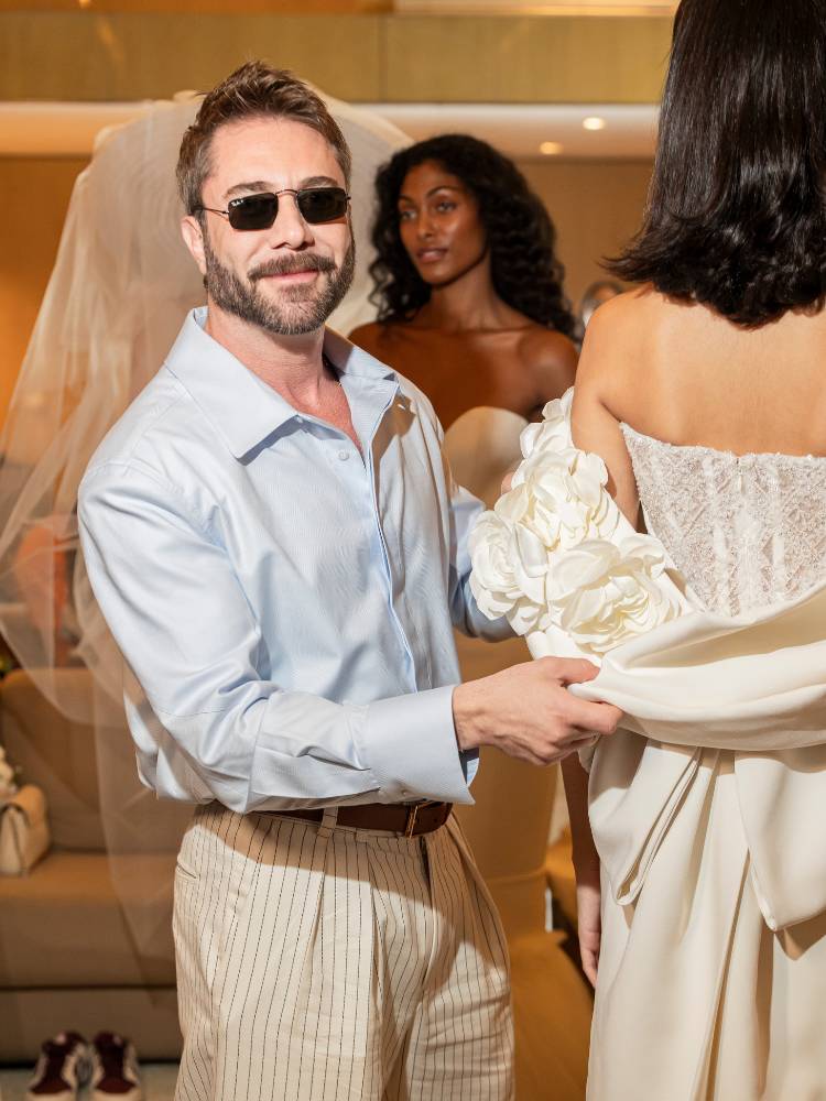 Lucas Anderi de óculos de sol, camisa azul, calça bege, mexendo em vestido de noiva