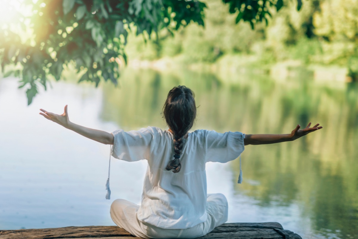 Mulher de braços abertos meditando em frente a um lago