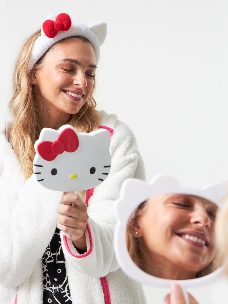Menina loira de pele clara usando pijama preto da Hello Kitty, com pijama de corpo todo da personagem, faixa da personagem e espelho da personagem