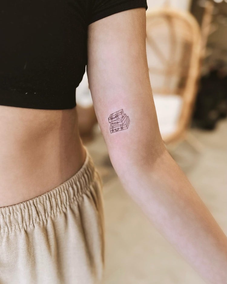 Tatuagem de traços de 3 malas uma em cima de outra em braço de mulher