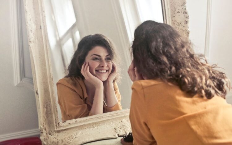 Foto de mulher morena de camisa amarela olhando a autoimagem no espelho