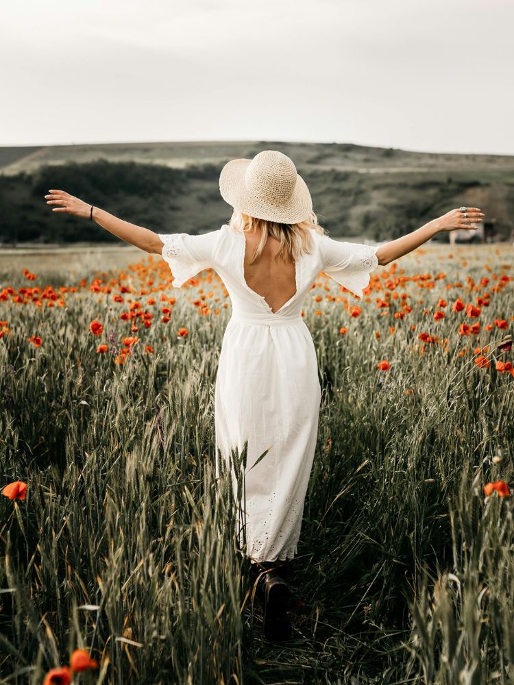 Foto de mulher de costas, com os braços abertos, em um campo de papoulas. Ela é branca, loira e usa chapéu de palha e vestido branco