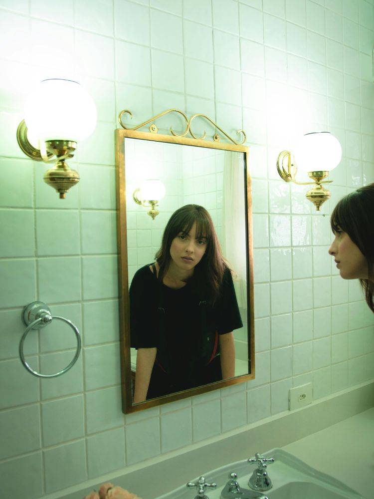 Foto de mulher de cabelo castanho longo com franja usando camiseta preta. Ela se olha no espelho em um banheiro de azulejos brancos