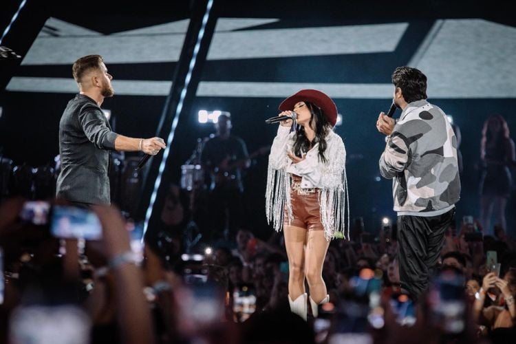 Foto da cantora Ana Castela usando chapéu de cowboy marrom, jaqueta branca de franjas e short de couro