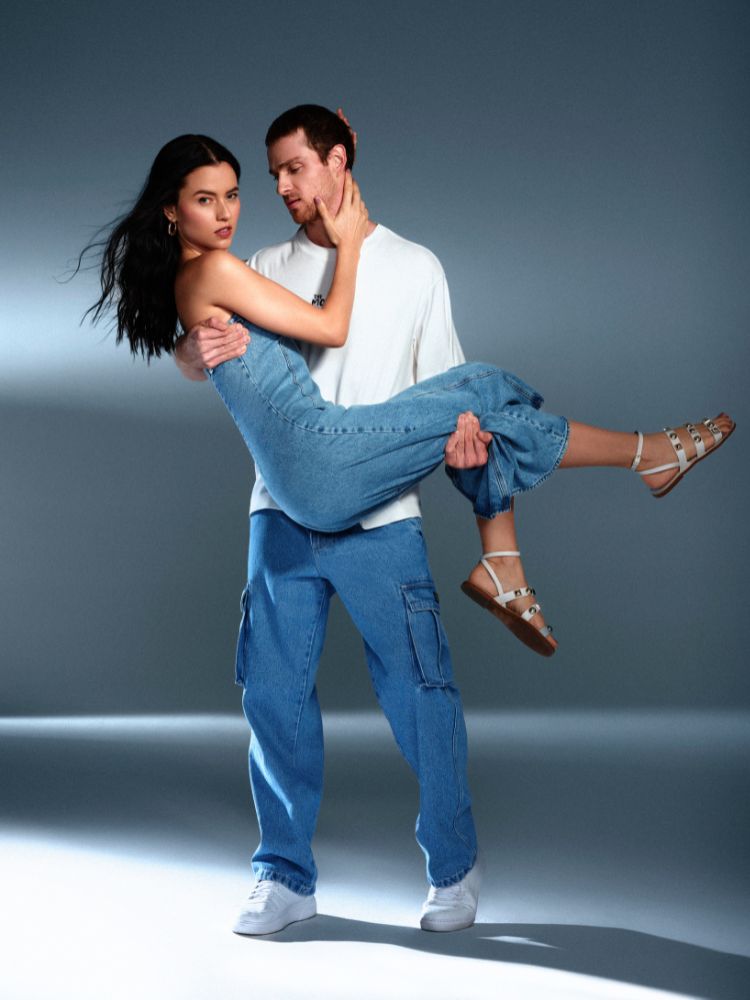 Foto de namorado carregando namorada no colo. Ele usa camiseta branca, calça cargo jeans e tênis, enquanto ela usa vestido jeans e rasteirinha 