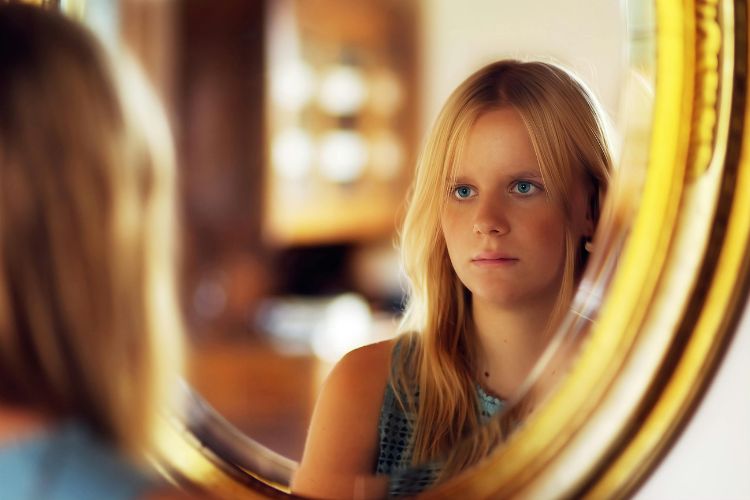 Foto de mulher loira de olhos azuis se olhando em espelho redondo de moldura dourada