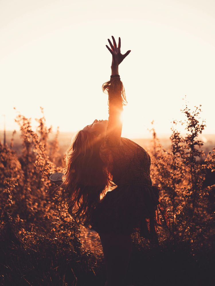 Foto de mulher de perfil, em meio à natureza no pôr do sol, com a mão direita estendida para cima