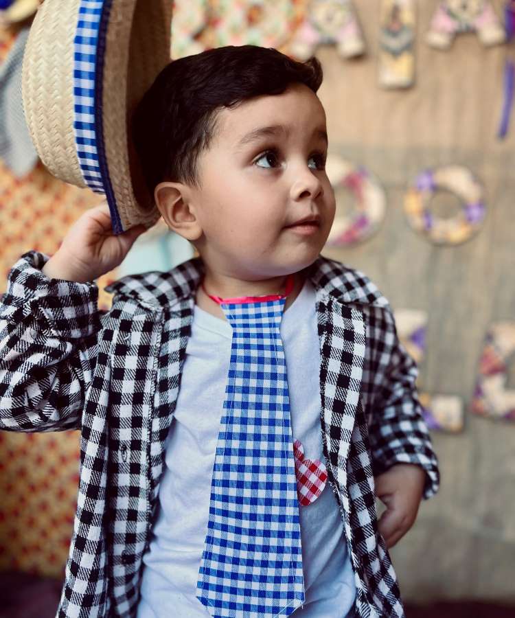 Menino usando camisa xadrez preta e branca, gravata xadrez azul e chapéu de palha.