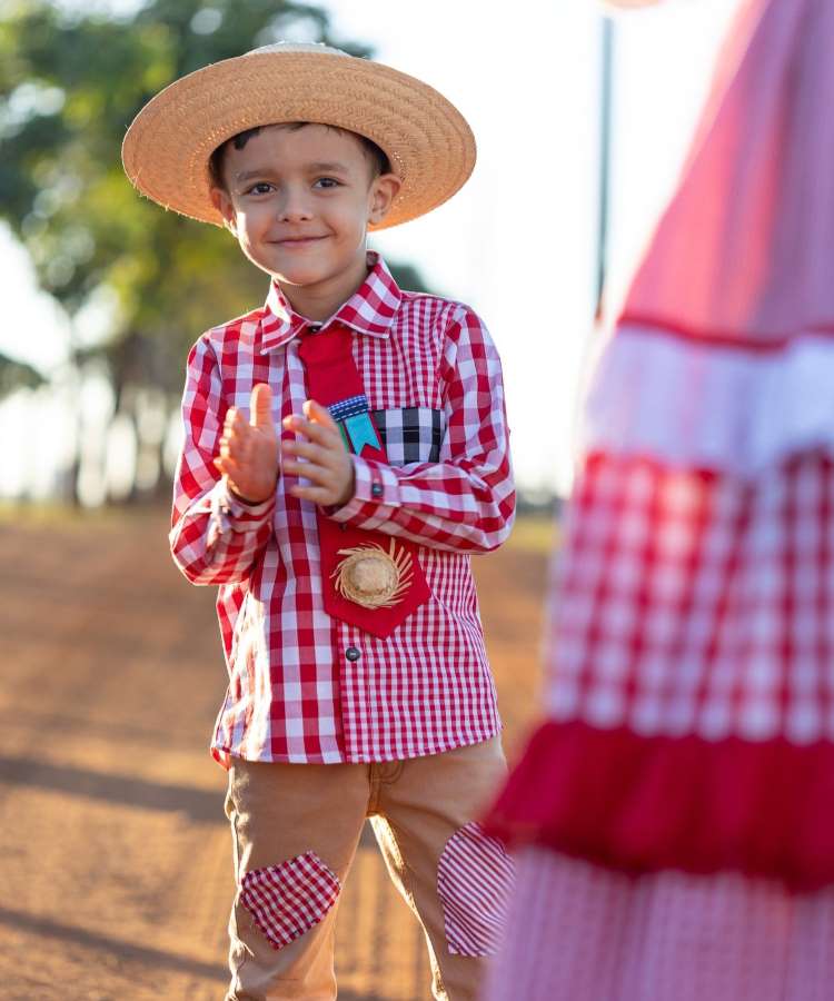 Menino usando camisa xadrez vermelha, calça jeans com detalhes e chapéu de palha para festa junina.