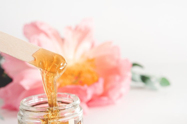 Pote de vidro com cera e palito com cera em fundo com flor rosa desfocado