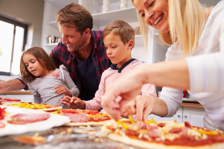 Mulher loira de pele clara, duas crianças de pele clara e homem de pele clara fazendo pizza com massa, molho de tomate, pimentão e salame
