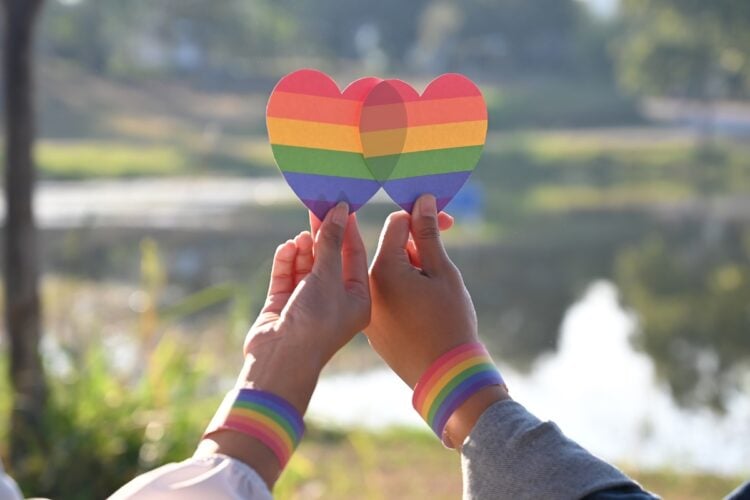 Foto de duas pessoas de pele clara, com pulseira colorida, ambas segurando coração com as cores da bandeira LGBT