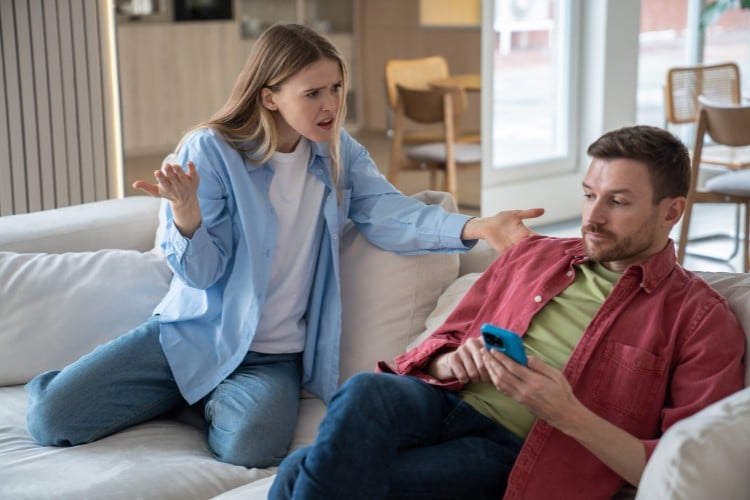 Mulher de pele clara usando blusa branca, camisa azul e calça jeans olhando indignada para homem de pele clara mexendo no celular, usando calça escura, blusa verde e camisa vermelha