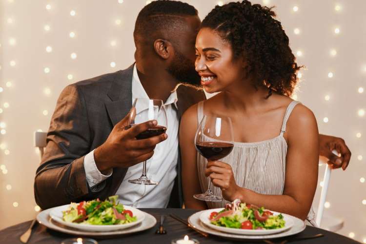 Casal em jantar romântico em casa, comendo salada e sgurando taças de vinho
