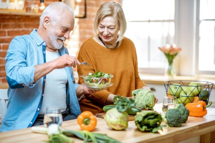 Homem idoso de blusa azul e mulher idosa de blusa laranja preparando alimentos saudáveis, com verduras e pimentão