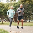 Mulher e homem idosos (ela de blusa azul e calça preta e ele de blusa cinza e bermuda preta) correndo em um parque