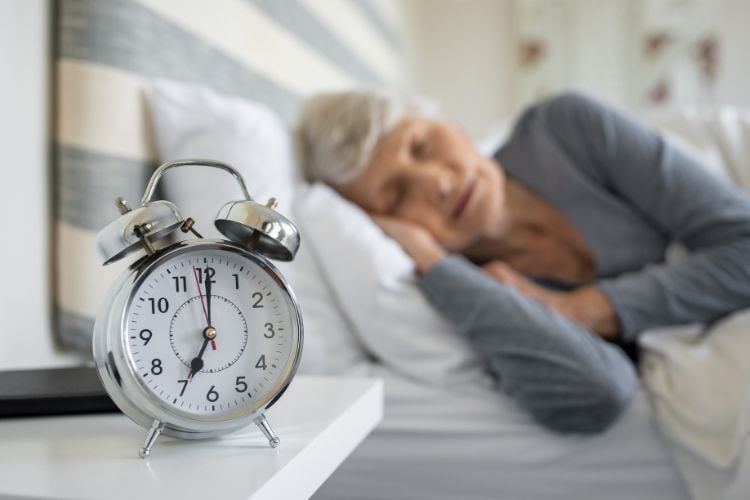 Mulher idosa de pele clara usando blusa cinza dormindo em cama ao lado de despertador