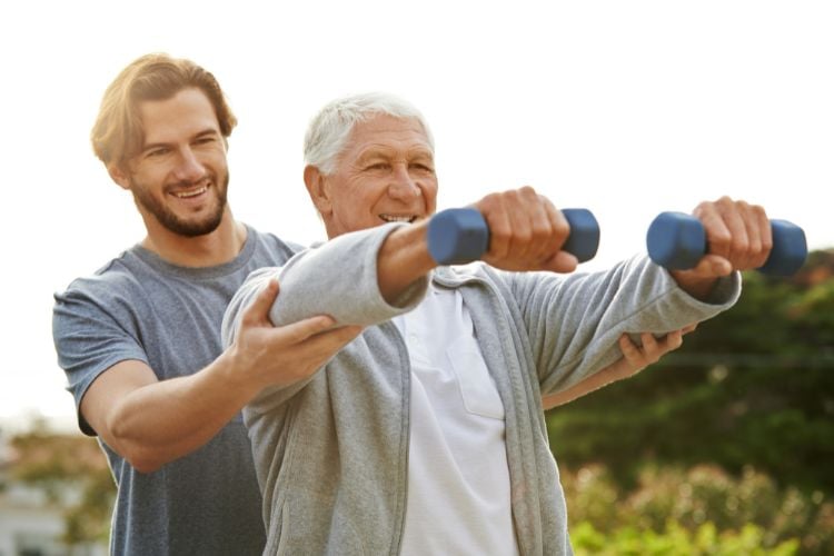 Homem jovem de blusa cinza ajudando homem idoso com exercícios de peso