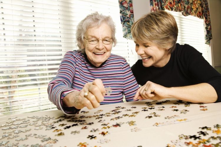 Mulher idosa de blusa roxa ao lado de mulher loira de pele clara usando blusa preta, ambas jogando quebra-cabeça