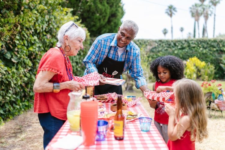 Mulher idosa de blusa vermelha e calça azul, homem negro idosos de blusa azul e avental preto e crianças ao redor de uma mesa com comidas e bebidas