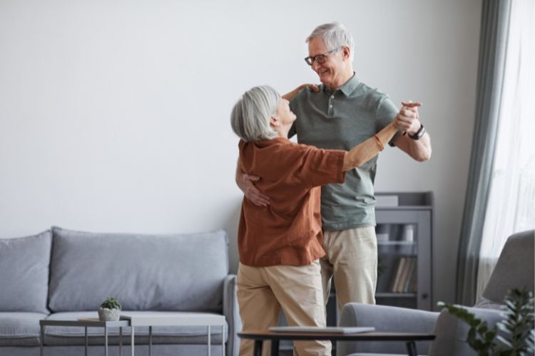 Homem e mulher idosos  (ela usando blusa marrom e calça bege e ele de blusa verde e calça bege) dançando em sala com sofá cinza