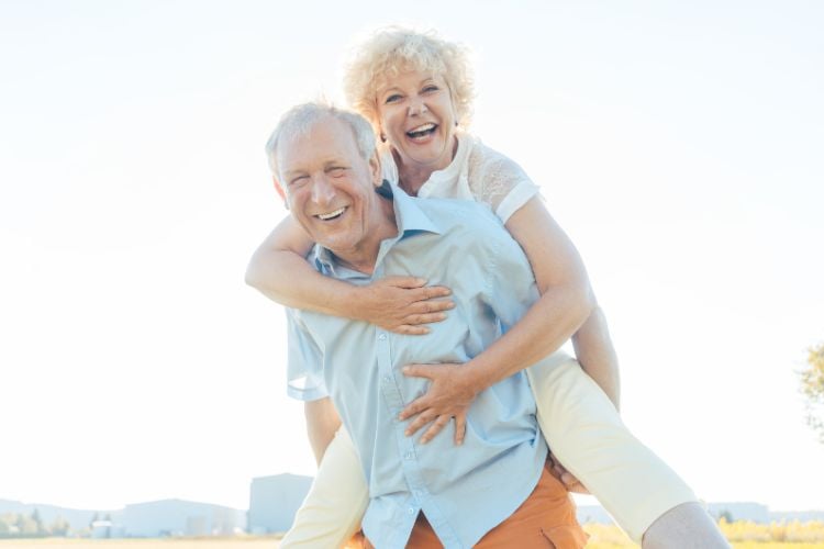 Homem idoso de camisa azul e calça laranja com mulher idosa de blusa branca em suas costas