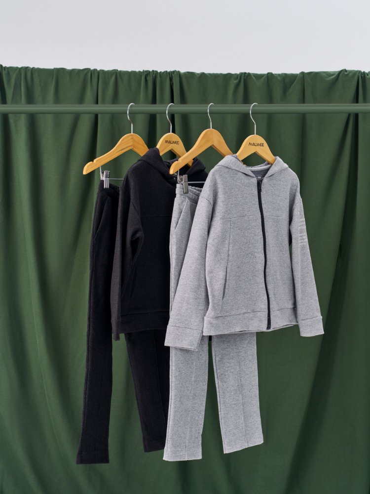 Foto de dois conjuntos de calça e jaqueta de moletom, um cinza e outro preto, pendurados em arara e na frente de pano verde