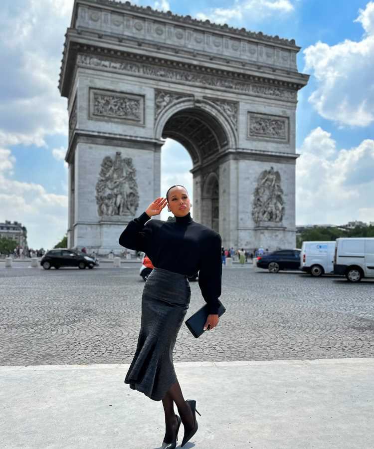 Kerry Washington usando Blusa preta, saia cinza, salto alto preto e segurando um livro azul. Look elegante e casual, ideal para passeios, na Semana de Alta Costura de Paris inverno 2024
